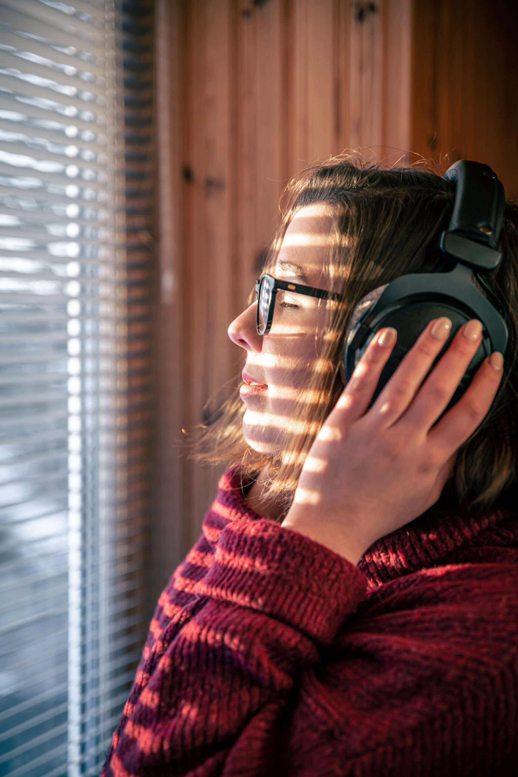 A young woman enjoys listening to music on headphones early in the morning in the sunlight through the blinds on the window, beautiful shadows.