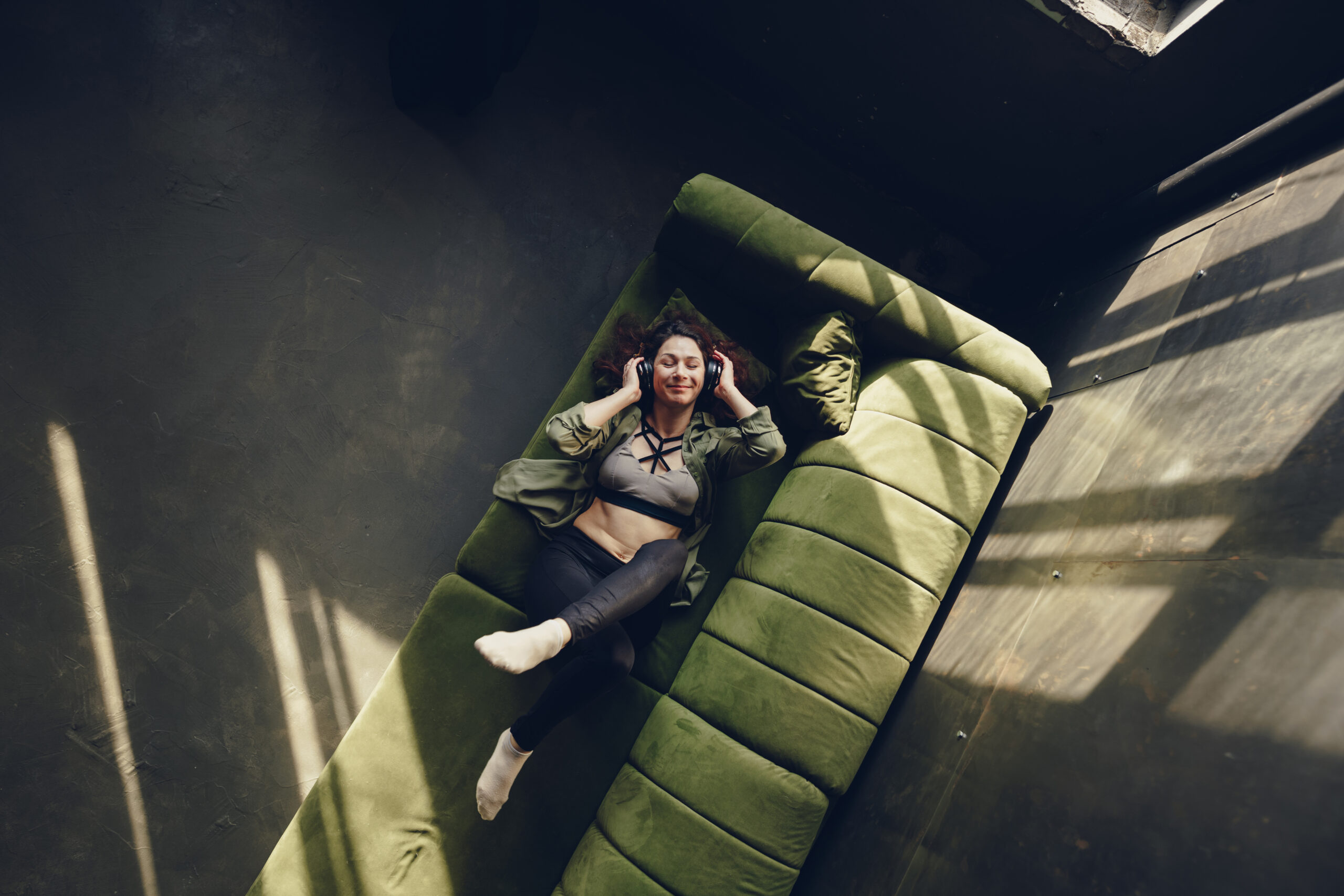 Brunette woman with a smile in headphones with closed eyes on the sofa, top view. An optimistic middle-aged woman listens to a positive melody lying on the sofa with her legs crossed. Top view, copy space