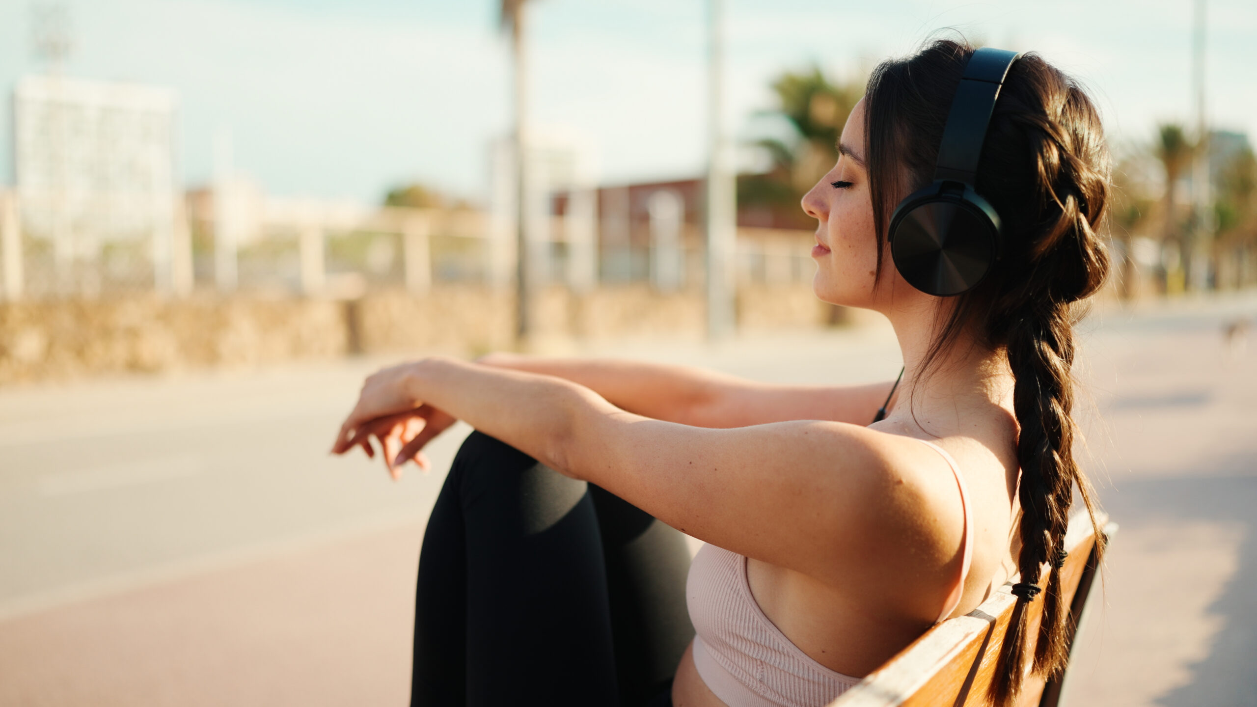Close up of young athletic woman with braided pigtail wearing beige sports top with headphones sits on bench relaxing and resting on the sea background. Beautiful girl enjoying music