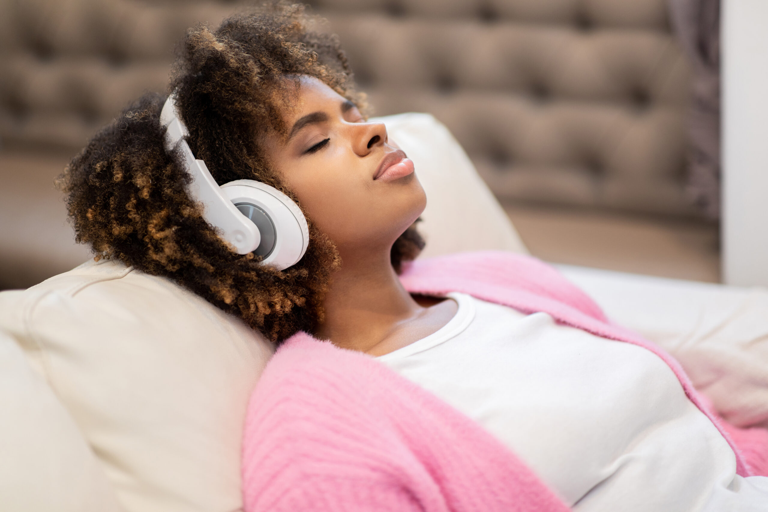Closeup of peaceful relaxed millennial black woman with bushy hair wearing comfy casual outfit chilling on couch with closed eyes at home, using wireless headphones, copy space