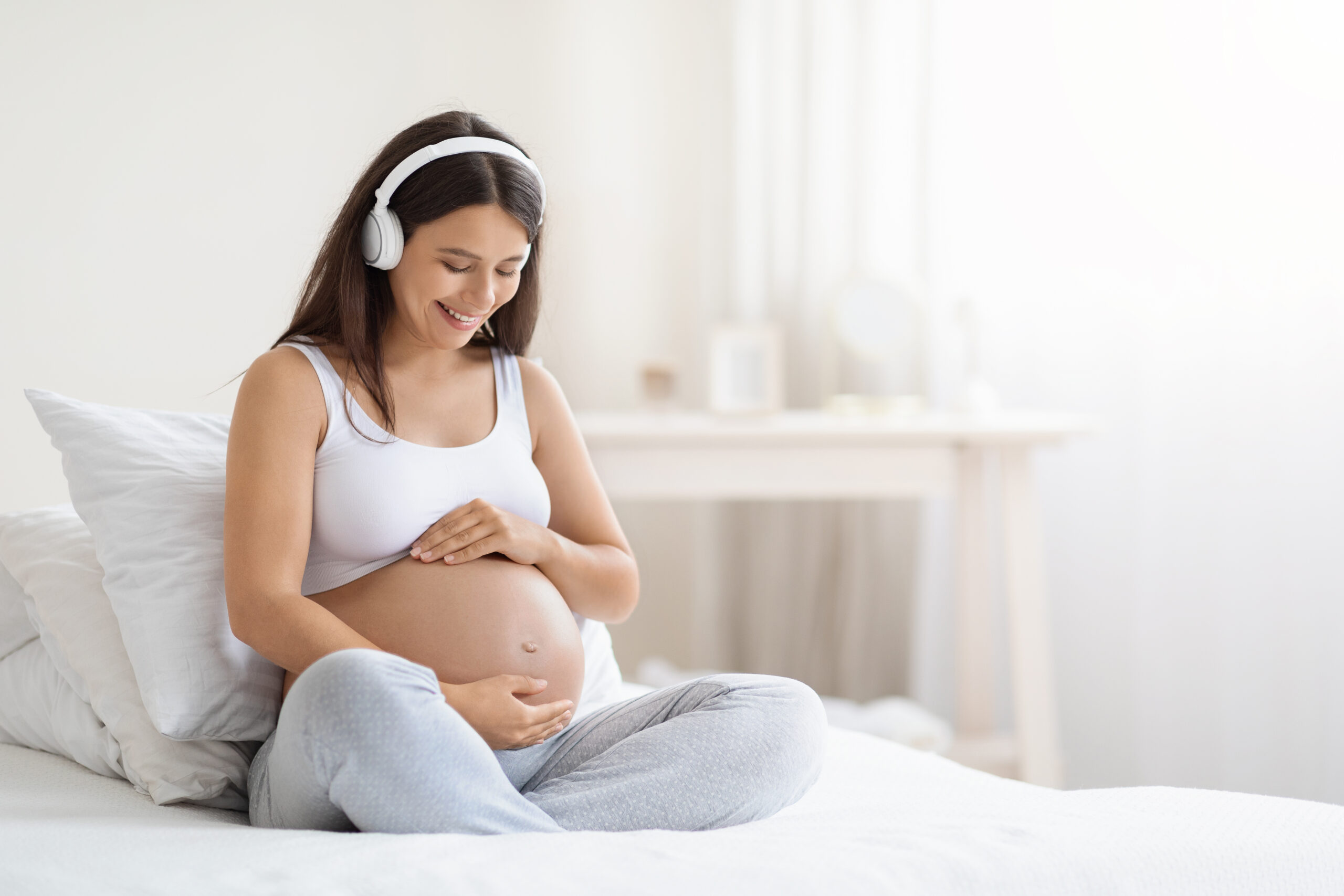 Happy smiling young pregnant woman relaxing, listening to music while sitting on bed at home, touching her big tummy, white bedroom interior, panorama with copy space. Music for baby in womb concept