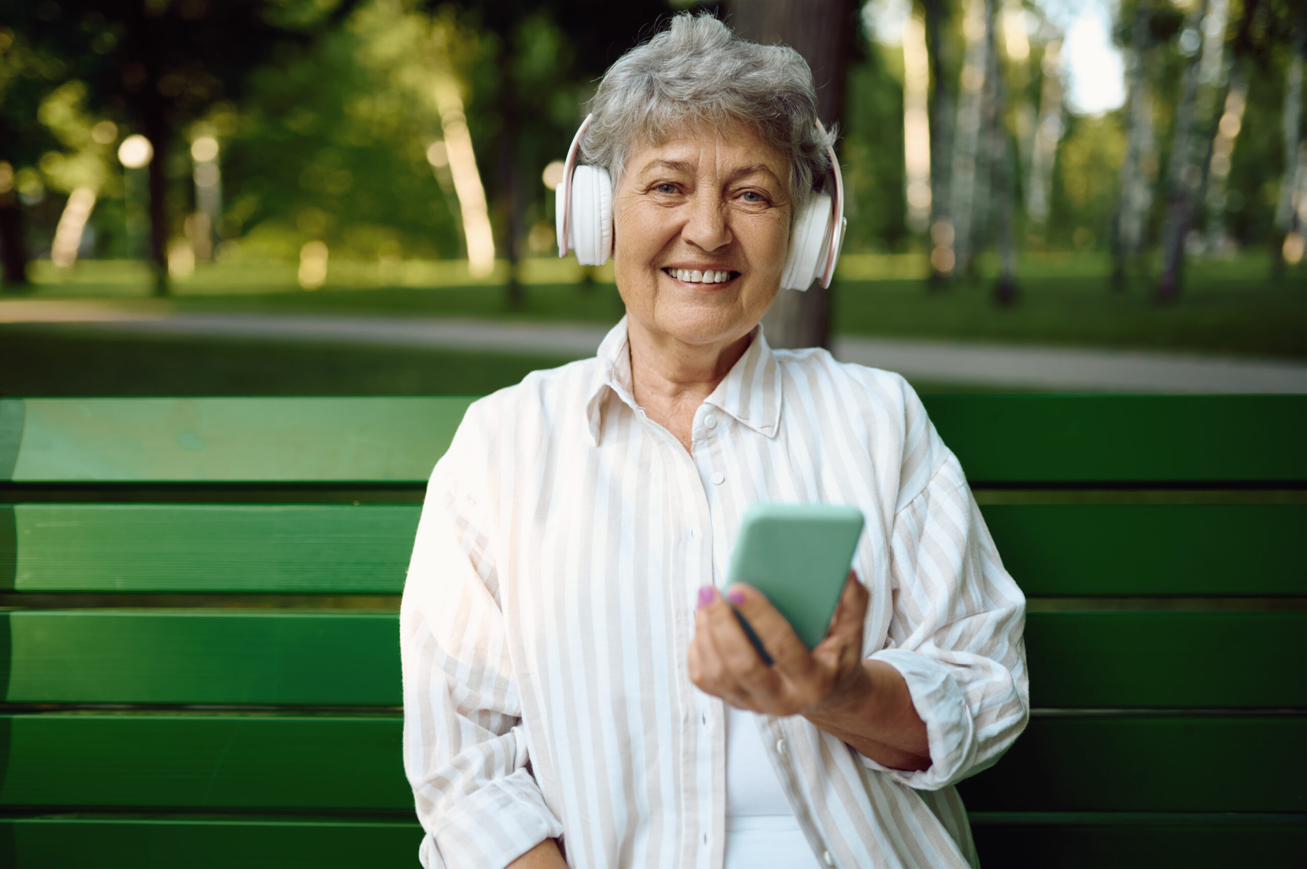 Old woman in headphones listens to music on the bench in summer park. Aged people lifestyle. Pretty grandmother having fun outdoors, an elderly female person on nature