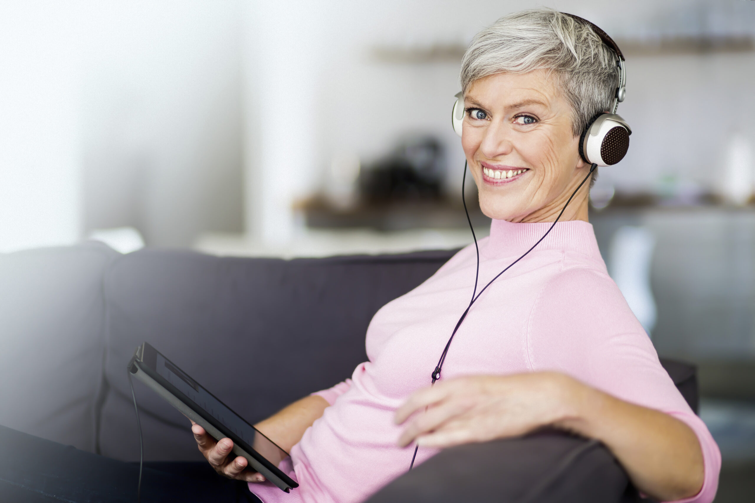 Portrait of happy mature woman sitting on couch with digital tablet listening music with headphones